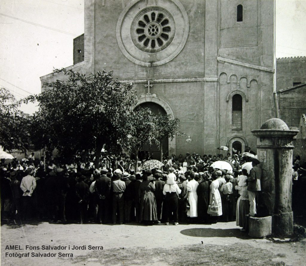 Cercle de persones davant l’església amb motiu d’una ballada. En primer terme, la font que hi havia a la plaça. Dècada de 1920.